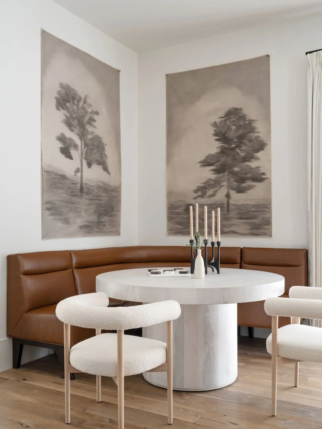 A dining room with a round white marble table, a curved brown leather banquette, two cream upholstered chairs, and two black candle holders with white candles on the table. Two large black and white landscape art prints of trees are hanging on the white walls.