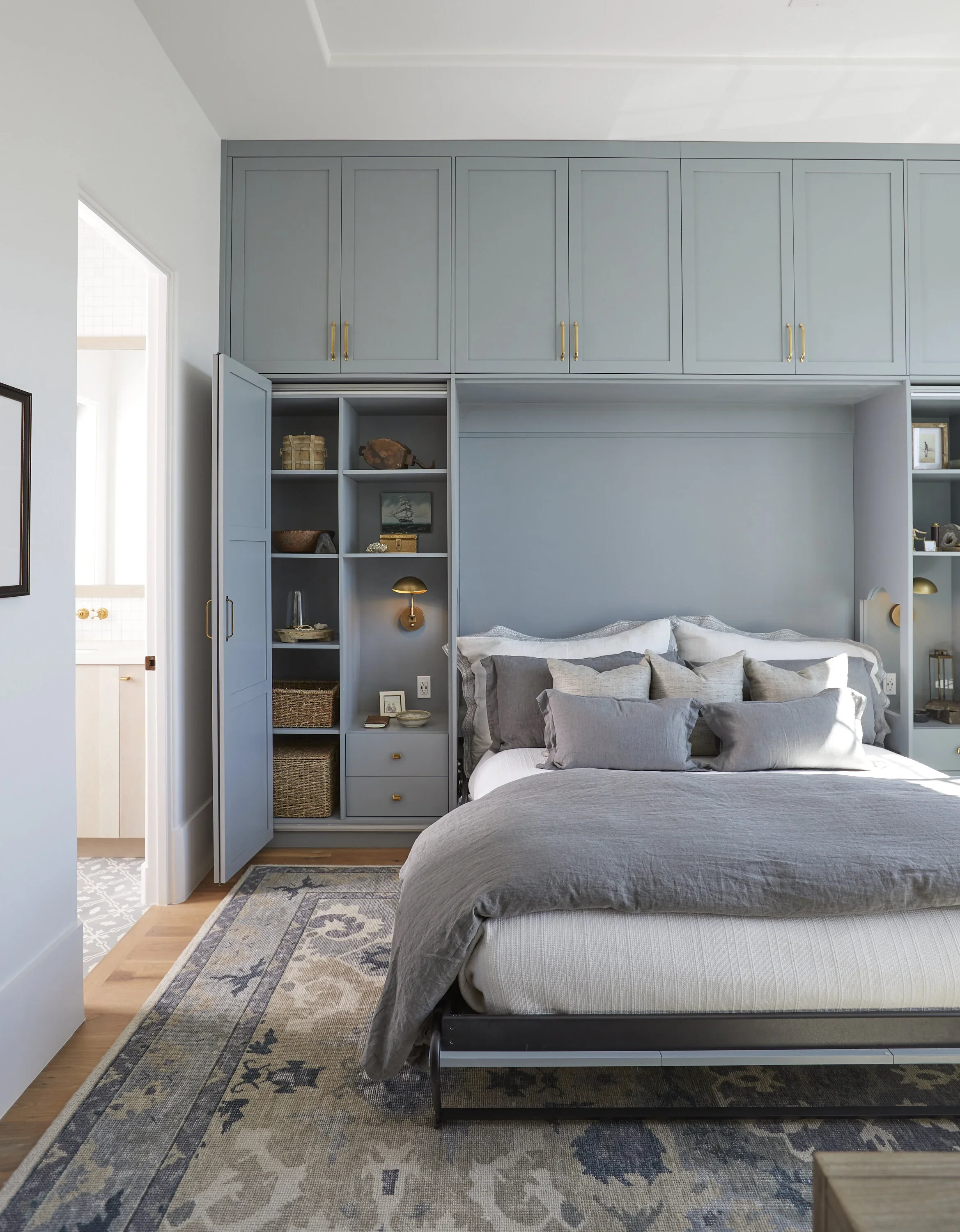 A modern bedroom with a gray bed, multiple gray pillows, and a built-in storage wall with upper cabinets and open shelves decorated with books, art, and decorative items. A patterned rug is on the wooden floor.