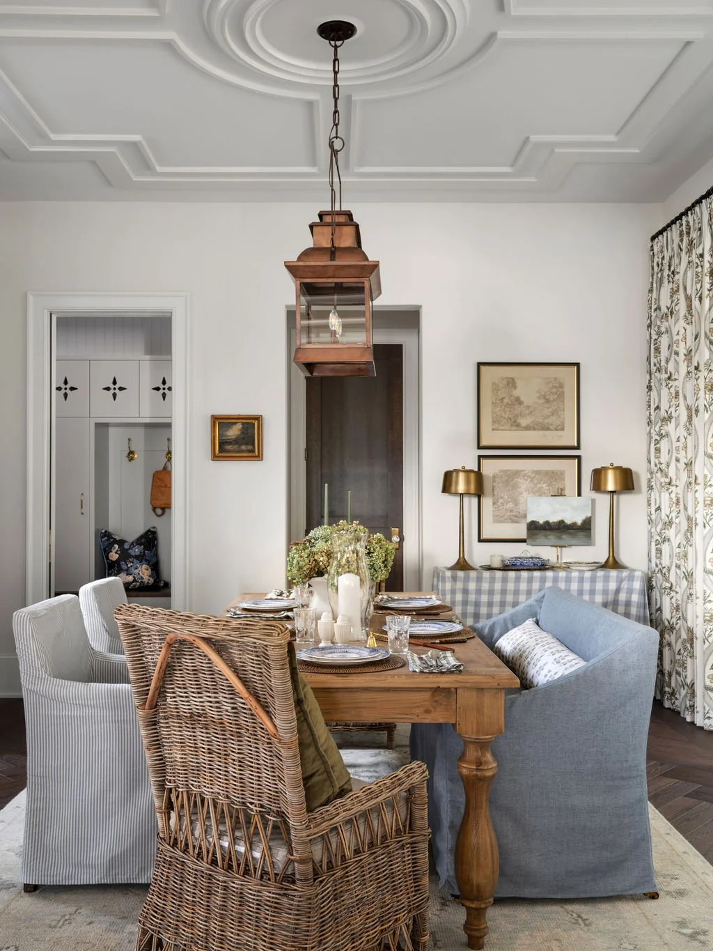 A dining room with a wooden table set for a meal, surrounded by a mix of woven and upholstered chairs, a blue sofa, and decorated with framed artwork, gold lamps, and a floral curtain.
