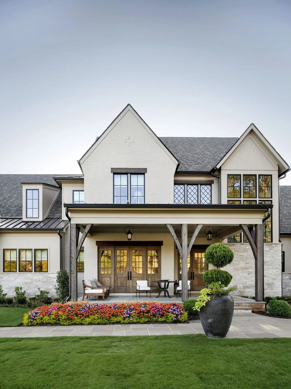 Front view of a modern house with a covered porch, potted plant, green lawn, and colorful flower bed.