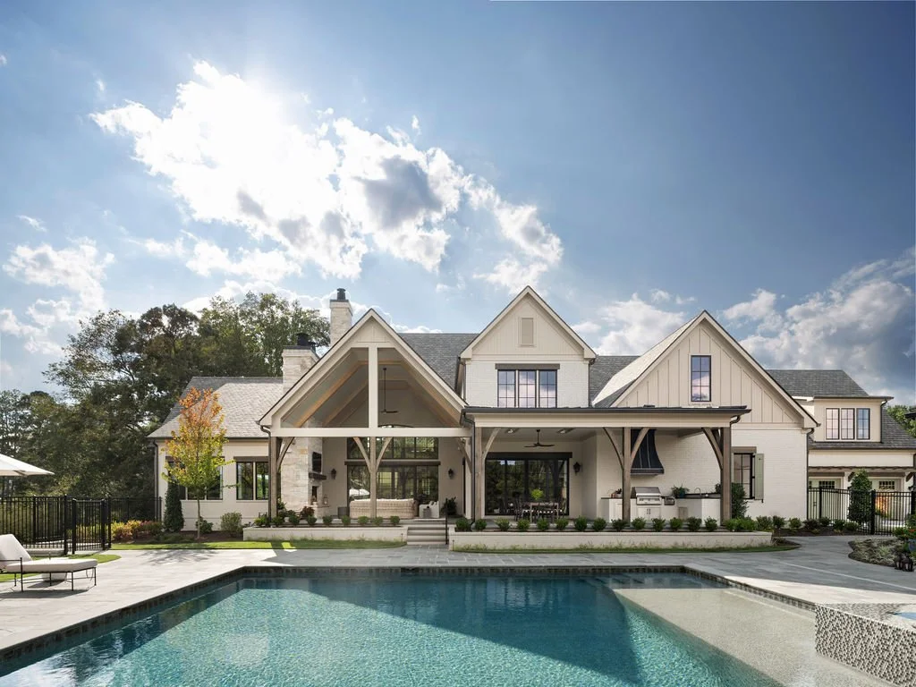 Large modern house with a backyard pool, patio area, and outdoor kitchen, featuring a gabled roof, white exterior, and surrounding greenery.