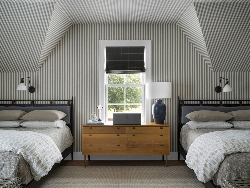 A bedroom with two beds, patterned striped walls, a window with a black shade, a wooden dresser with a lamp and other items, and black wall-mounted reading lamps.
