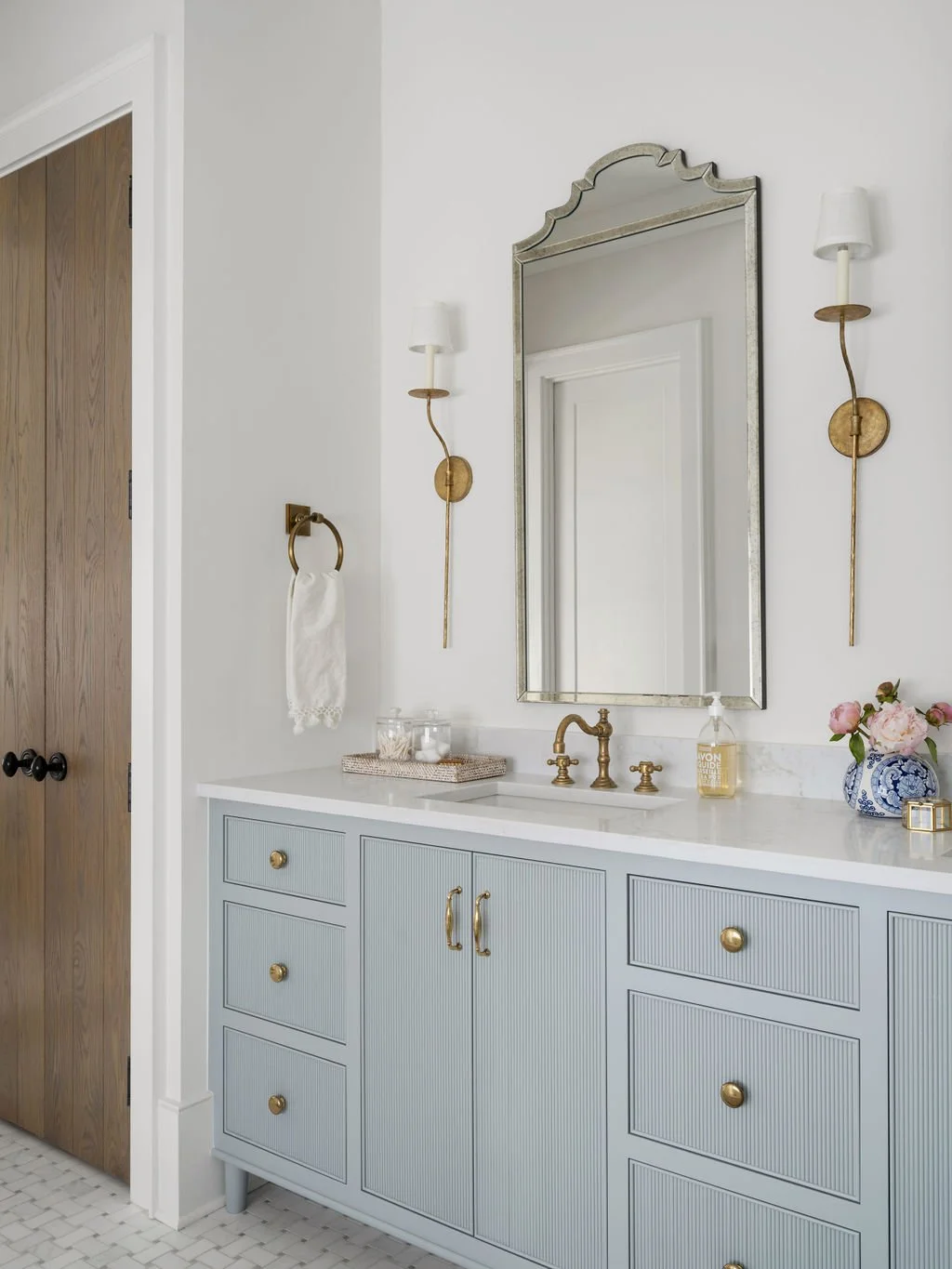 Bathroom vanity with a light blue cabinet, white countertop, a brass faucet, and a large mirror. Flanked by two gold wall sconces with white shades. Decor includes a clear jar, soap dispenser, and a blue and white vase with pink flowers.