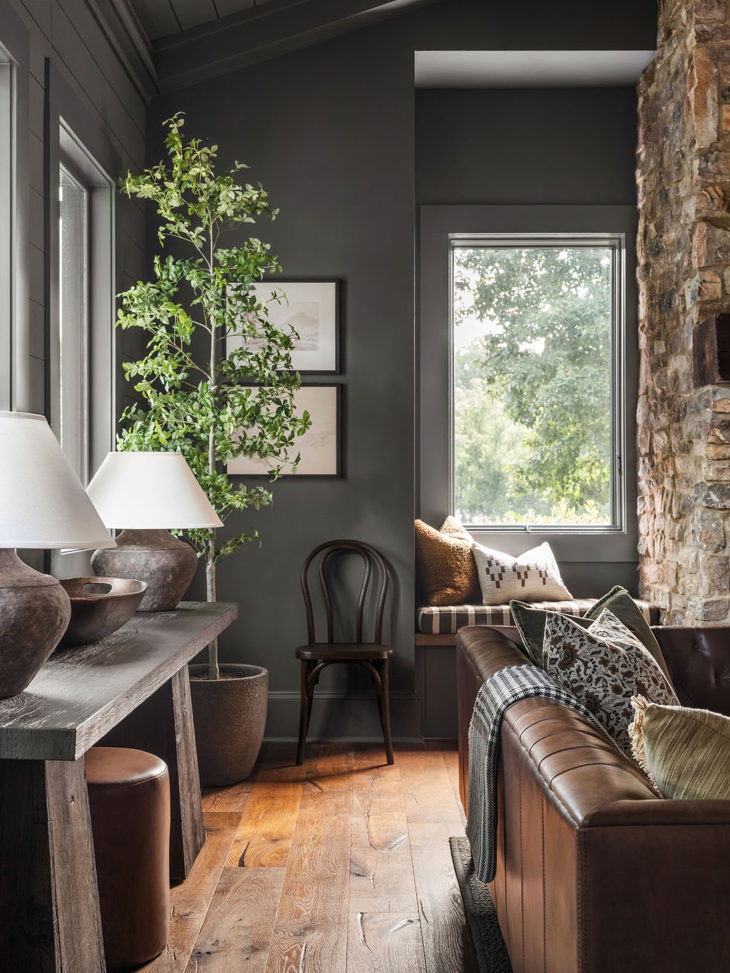 Interior of a cozy living room with dark walls, wooden flooring, a stone wall, a window with natural light, a leather sofa with cushions, a wooden chair, a large green plant, framed artwork, and decorative lamps.