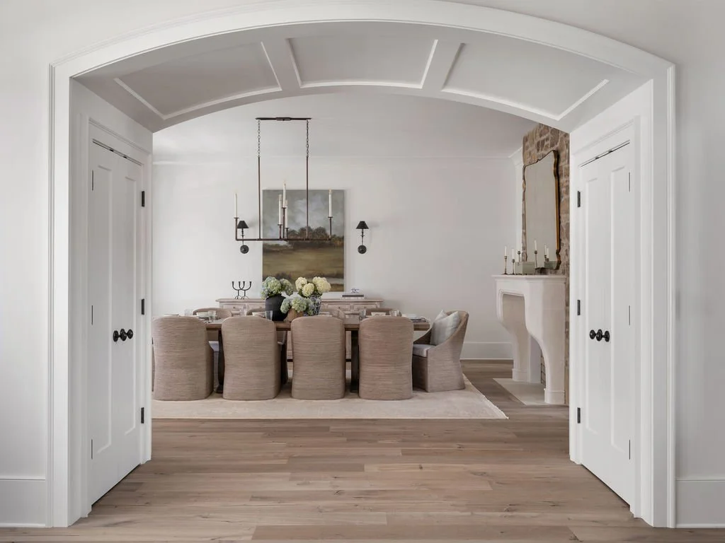 A dining room viewed through white double doors with black knobs, featuring a wooden table, beige upholstered chairs, a chandelier, a bouquet of white flowers, and wall-mounted lamps, with a fireplace and mirror on the wall.