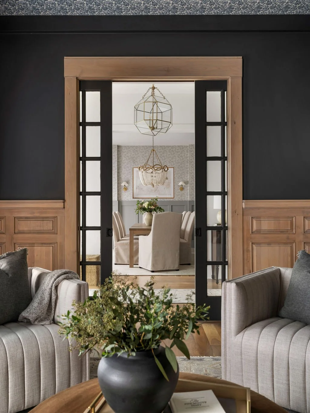 Living room with beige sofas and a coffee table with a black vase filled with greenery. A doorway with black-framed glass doors leads to a dining area with a round table, beige upholstered chairs, a chandelier, and a wall mirror.