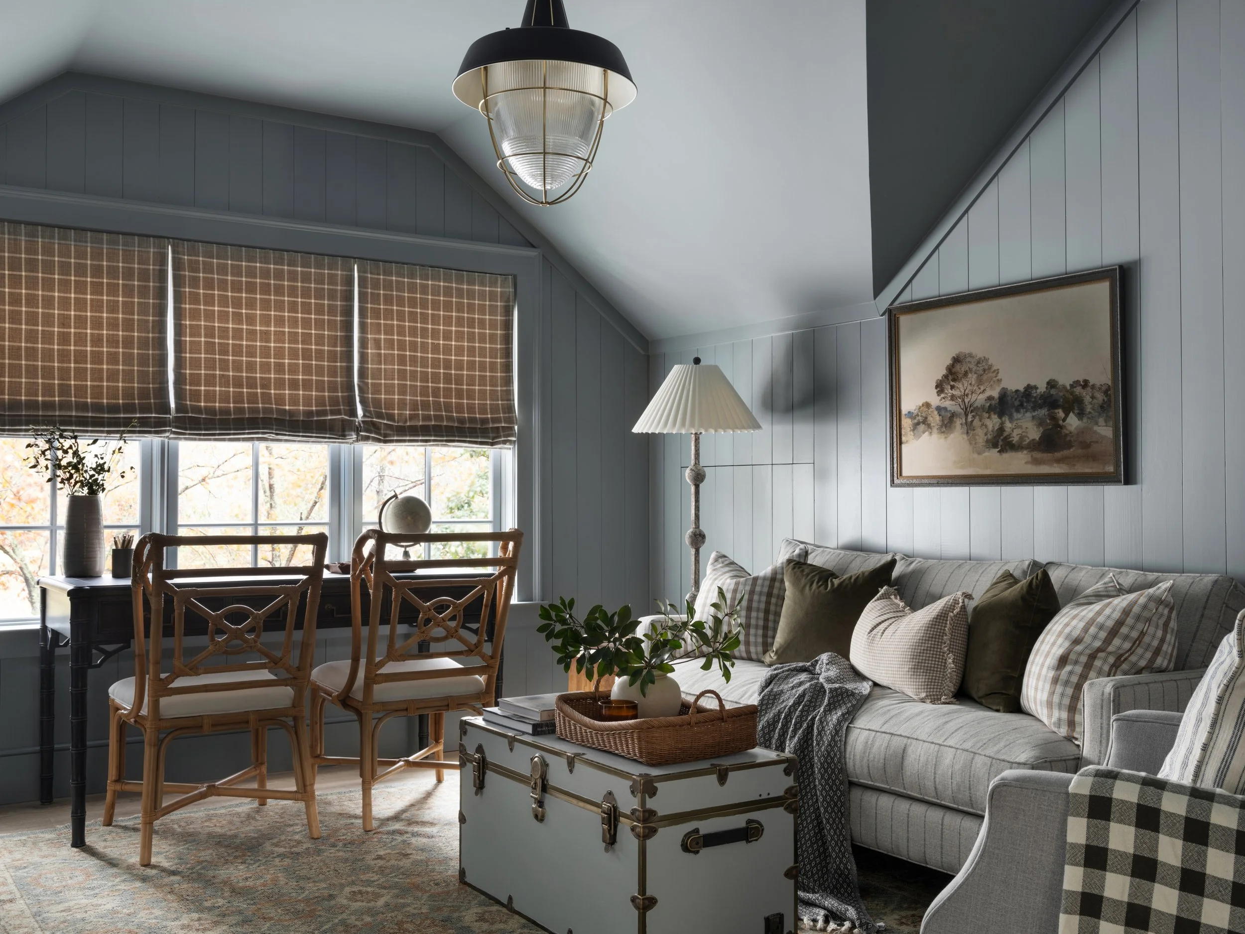 Cozy living room with a large gray sofa, multiple pillows, a white trunk used as a coffee table, and a floor lamp. A window with brown checkered shades, a small desk with a chair, and a framed painting of trees on the wall.