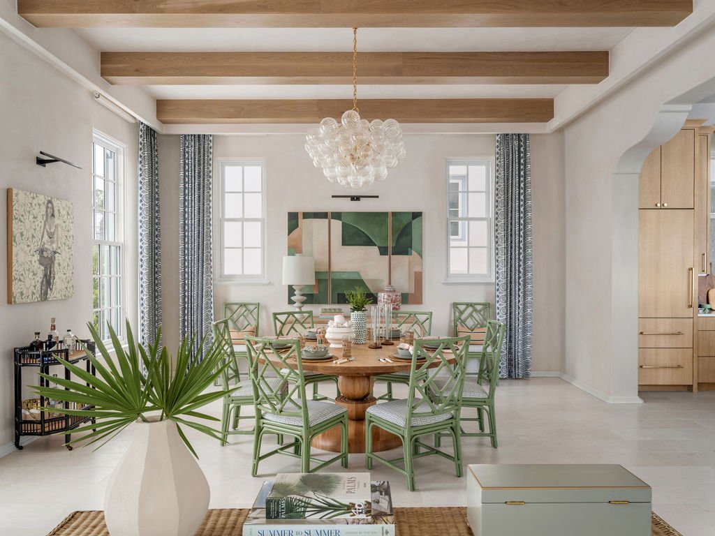 A modern dining room with a round wooden table, eight green chairs, a white vase with palm leaves, and artwork on the wall. It has wooden ceiling beams, a glass chandelier, two windows with patterned curtains, and a side bar with bottles.