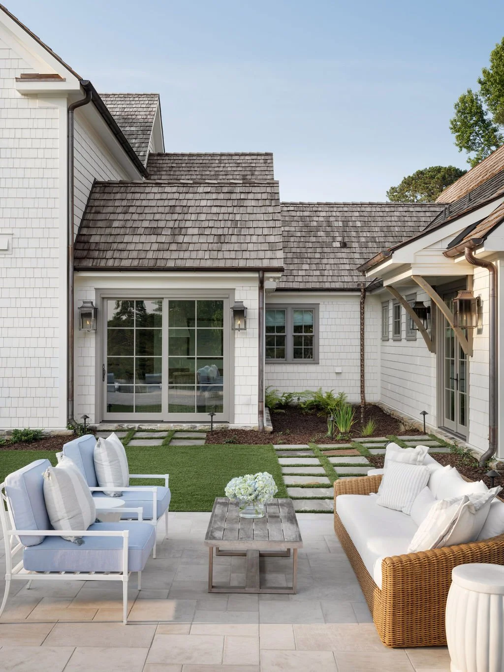 Outdoor patio area with white and beige seating, a wooden coffee table with flowers, and a well-maintained lawn with stepping stones leading to a house with white siding and a slide door.