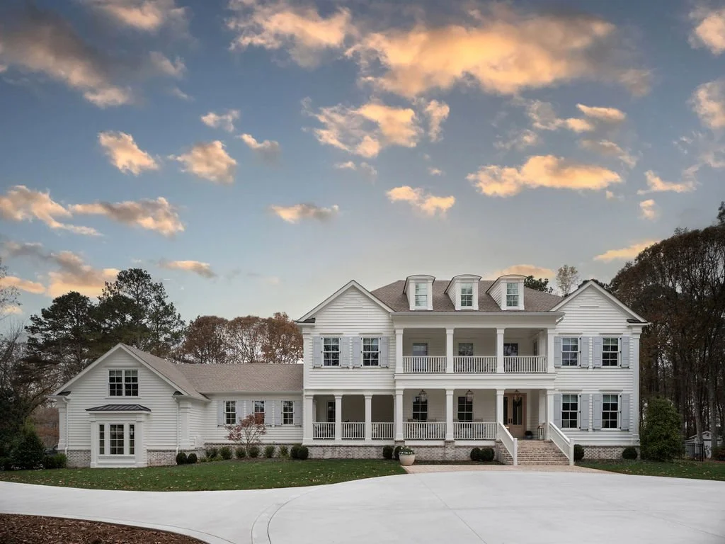A large white two-story house with a porch and dormer windows, set against a background of trees and a partly cloudy sky at sunset.