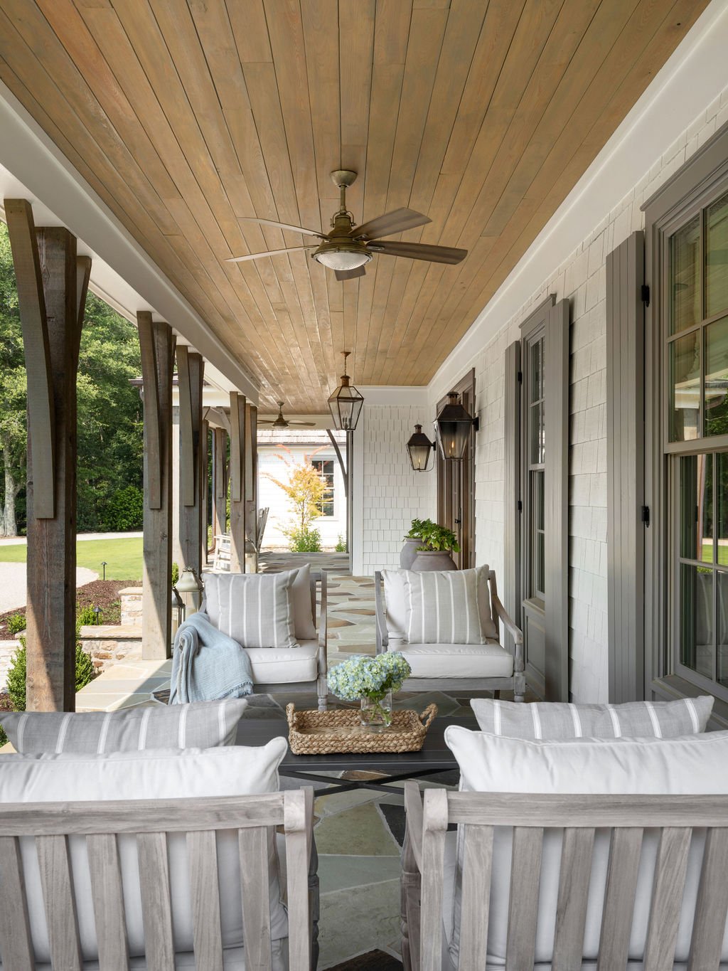 Outdoor porch with white and gray furniture, striped pillows, potted plants, and a wooden ceiling with ceiling fans and lantern-style wall lights.