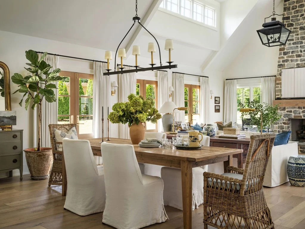 Bright and airy living and dining area with large windows, white curtains, a wooden dining table with white slipcovered chairs, a large vase with green flowers, a couch, wicker chairs, and decorative elements like lamps, plants, and artwork.