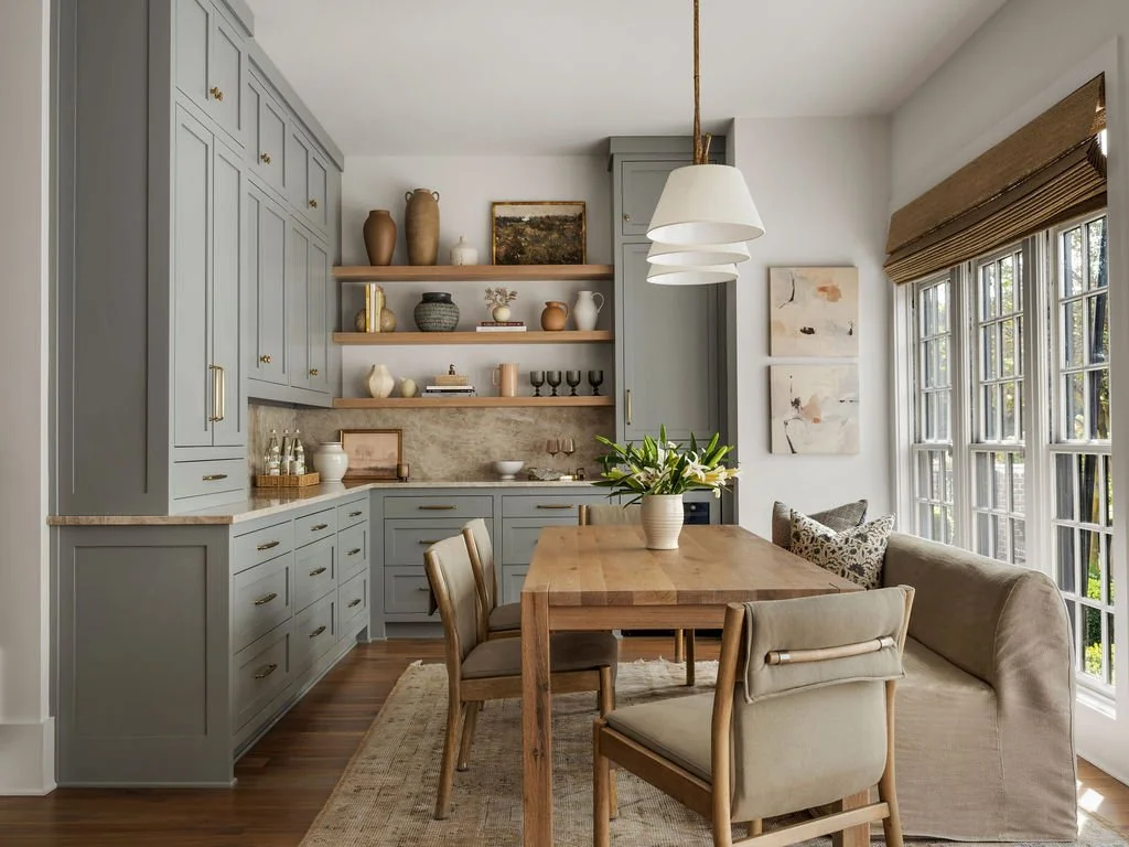A cozy dining area with a large wooden table, beige upholstered chairs, a beige sofa with patterned pillows, and a window with brown blinds. The kitchen features light blue cabinets, open wooden shelves with decorative items, and a pendant light hang