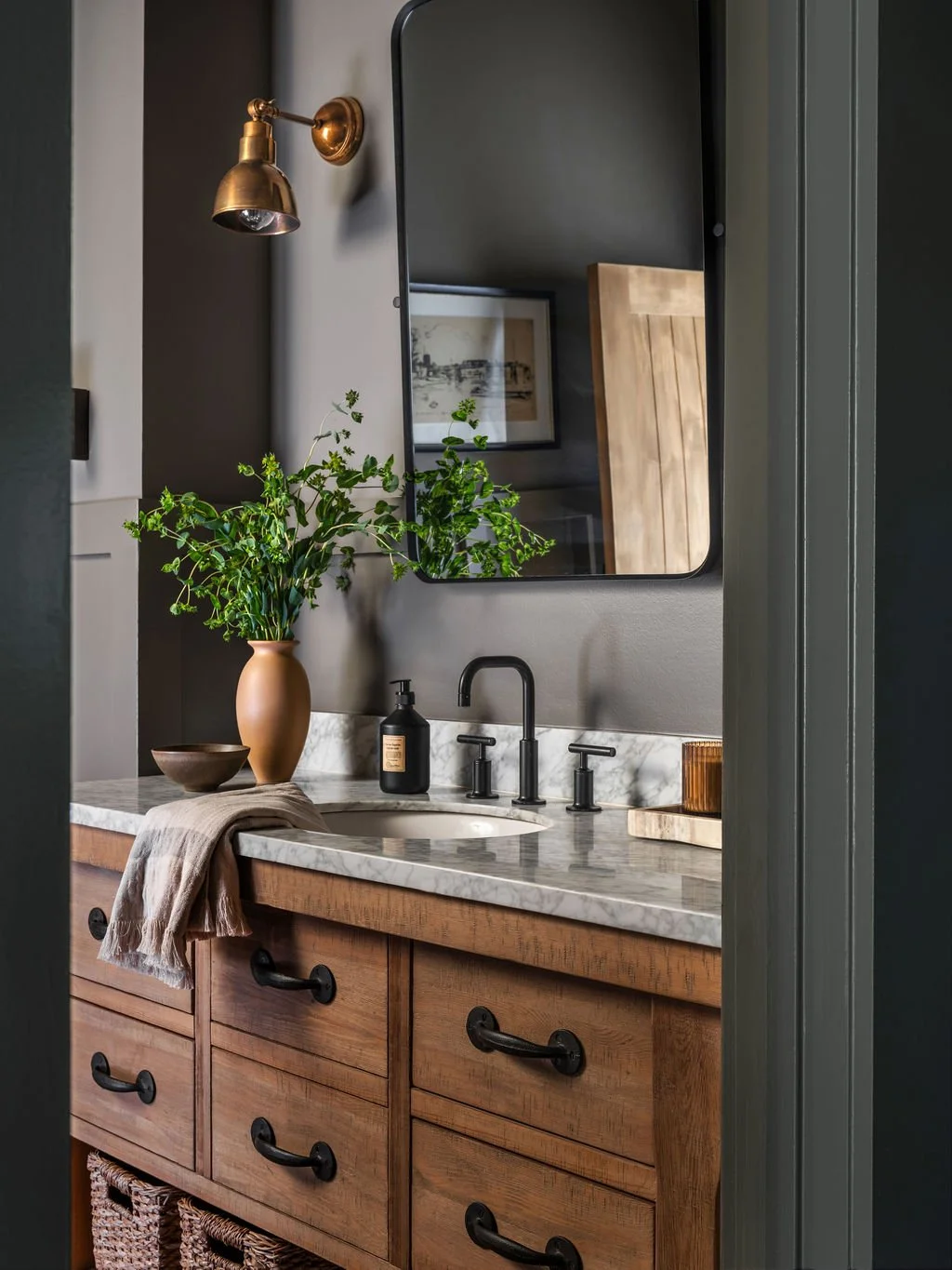 A bathroom vanity with a wooden cabinet, marble countertop, and black faucet. There is a mirror above, a potted green plant, a black soap dispenser, and a wooden tray. The wall lamp is mounted on the wall to the left of the mirror.