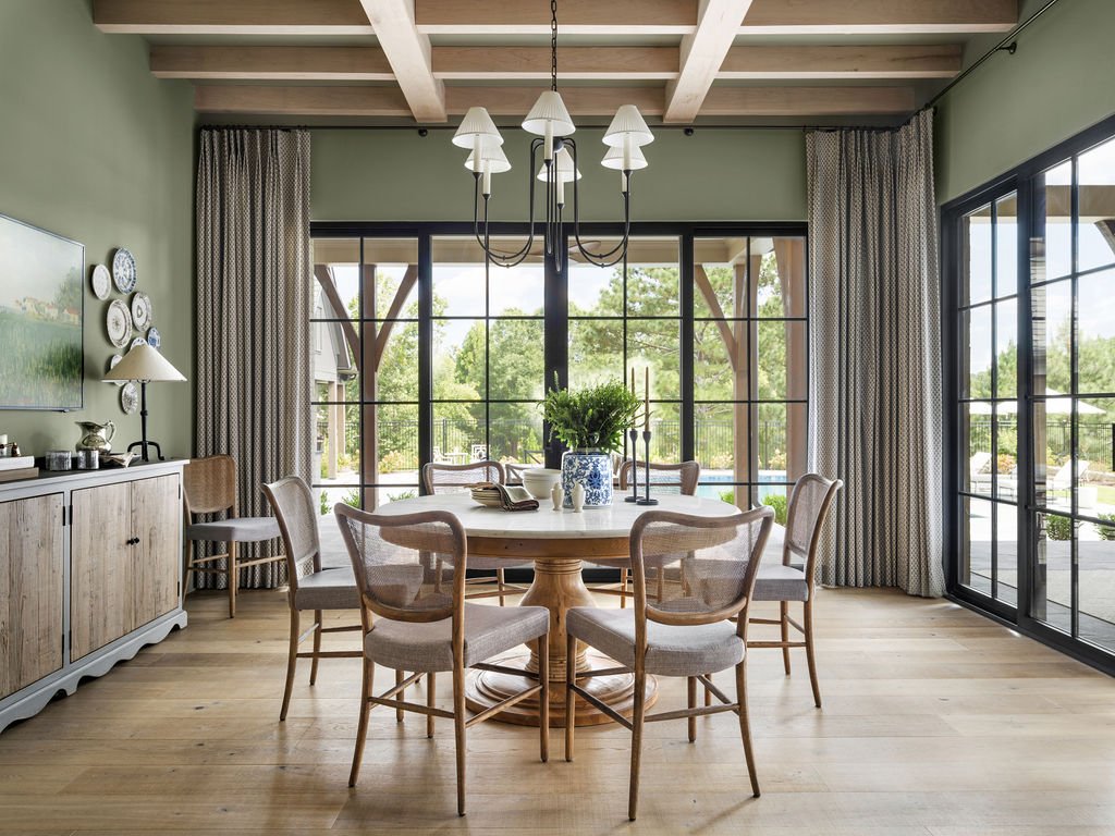 A dining room with a round wooden table surrounded by eight beige upholstered chairs. A chandelier hangs above the table. Large windows with patterned curtains allow natural light and offer a view of the outside patio and green trees.