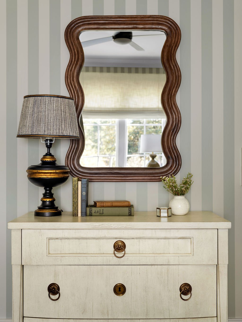 A white wooden dresser with brass handles, topped with a lamp, a small vase with plants, a stack of books, and a small decorative box, with a large brown wavy-edged mirror above it, reflecting a window and a ceiling fan.