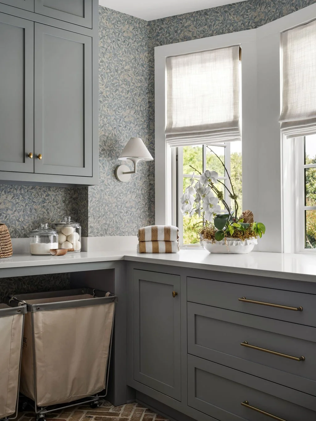 A cozy kitchen corner with light blue cabinetry, white countertops, floral wallpaper, white window shades, a bowl of orchids, and neatly folded towels.