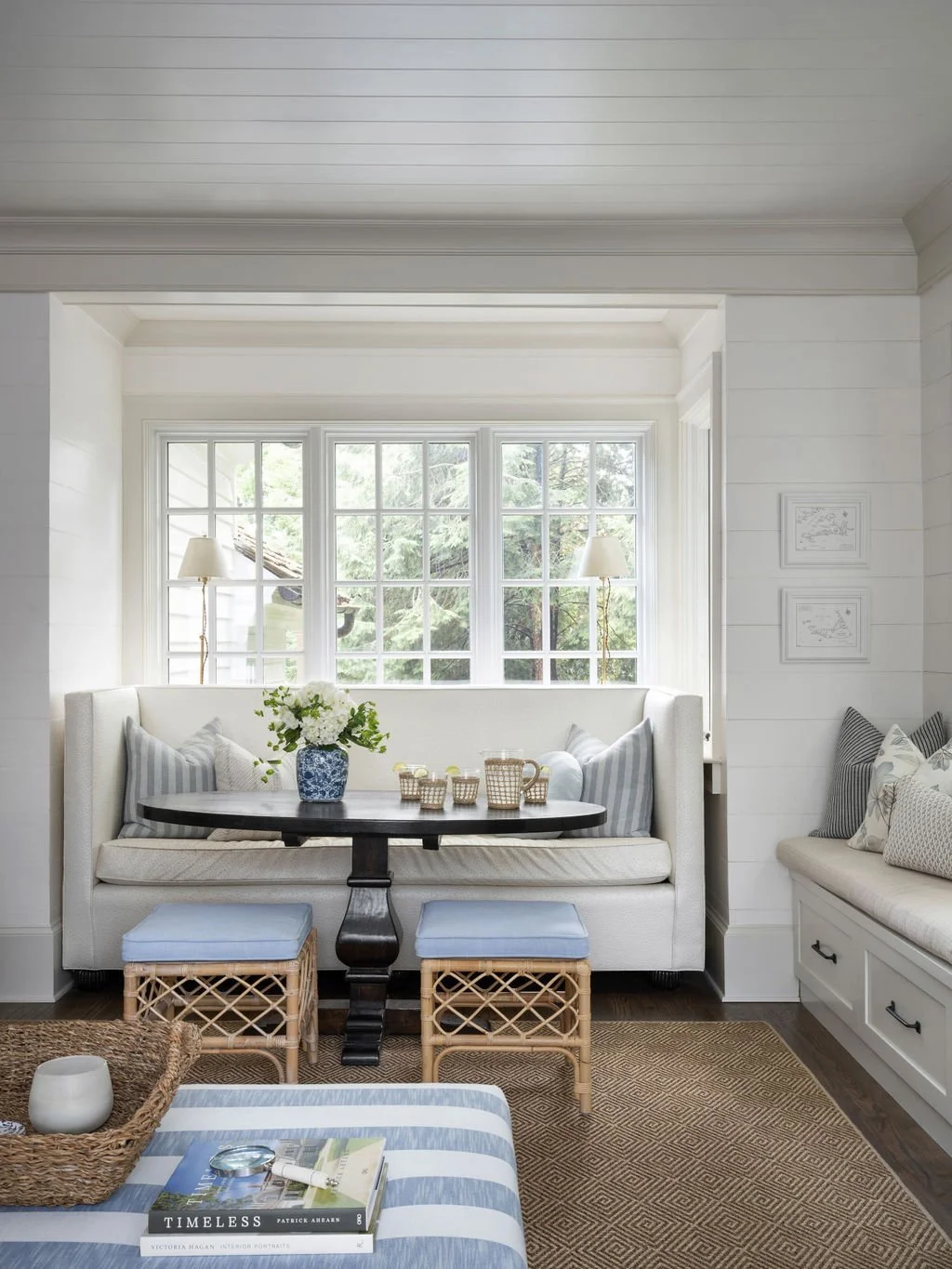 A cozy corner with a built-in white window seat, striped and patterned pillows, a dark wood table with wicker and blue stools underneath, and a window with greenery outside. A vase of flowers and cups sit on the table, with a basket, book, and decora