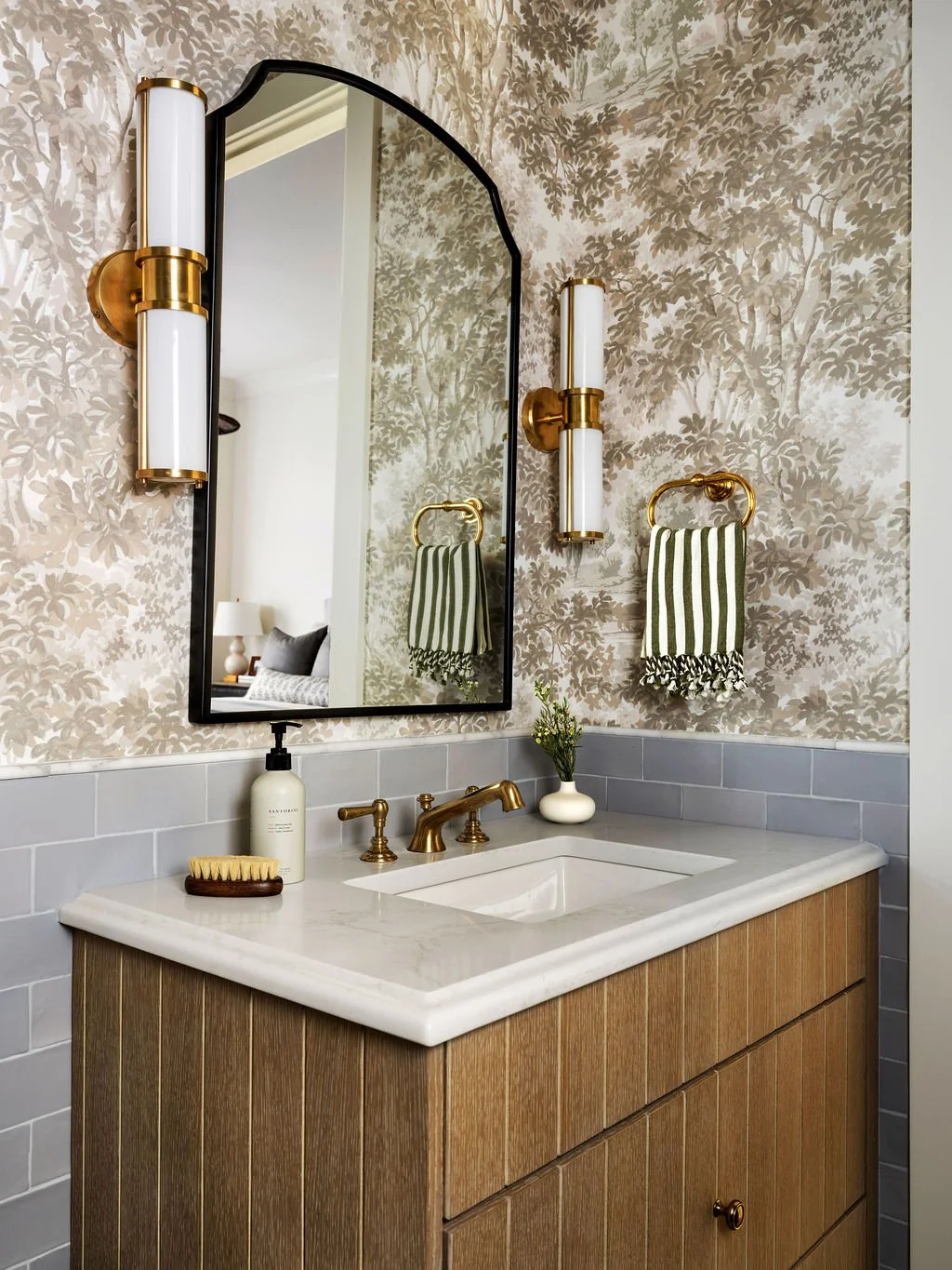 Bathroom vanity with white marble countertop, brass fixtures, a small vase with flowers, soap dispenser, and a wooden soap brush. Mirror with wall sconces and a striped towel on a brass towel ring.