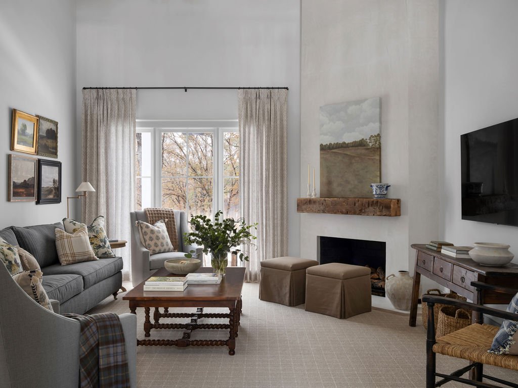 A cozy living room with a large window framed by curtains, a gray sofa with decorative pillows, a wooden coffee table with a plant, two ottomans, a TV on a wooden stand, and a fireplace with a wooden mantle.