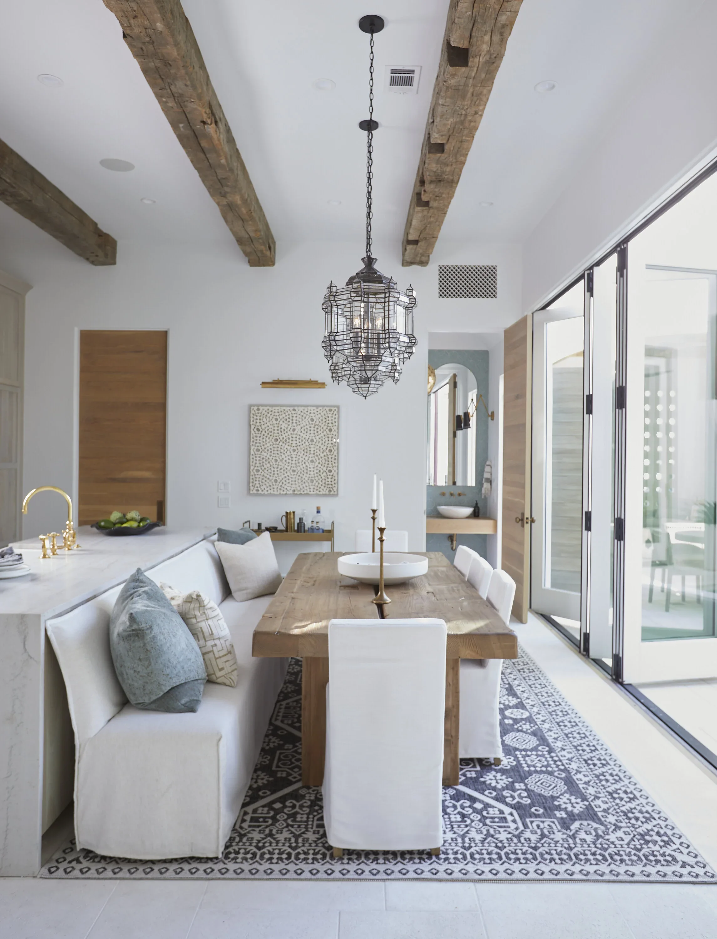 Dining area with a wooden table, white chairs, a patterned rug, large sliding glass doors, and a decorative overhead pendant light.