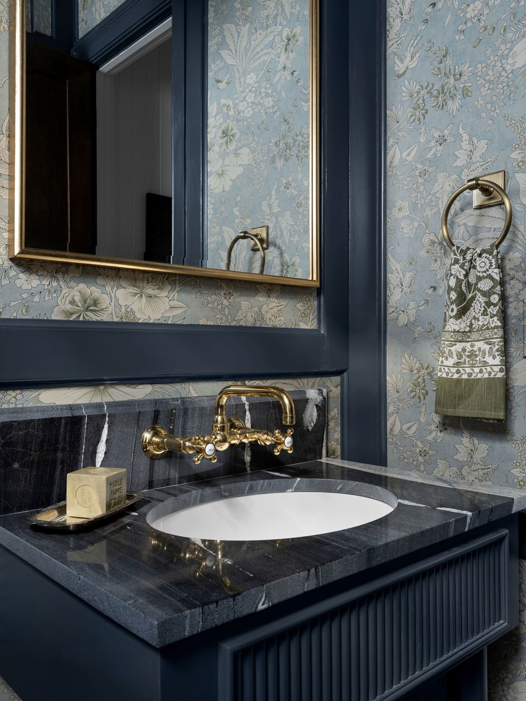 Bathroom vanity with a dark marble countertop, a white undermount sink, and gold fixtures. A mirror with a gold frame is above the sink, and a patterned towel hangs on a gold ring on floral wallpapered wall.