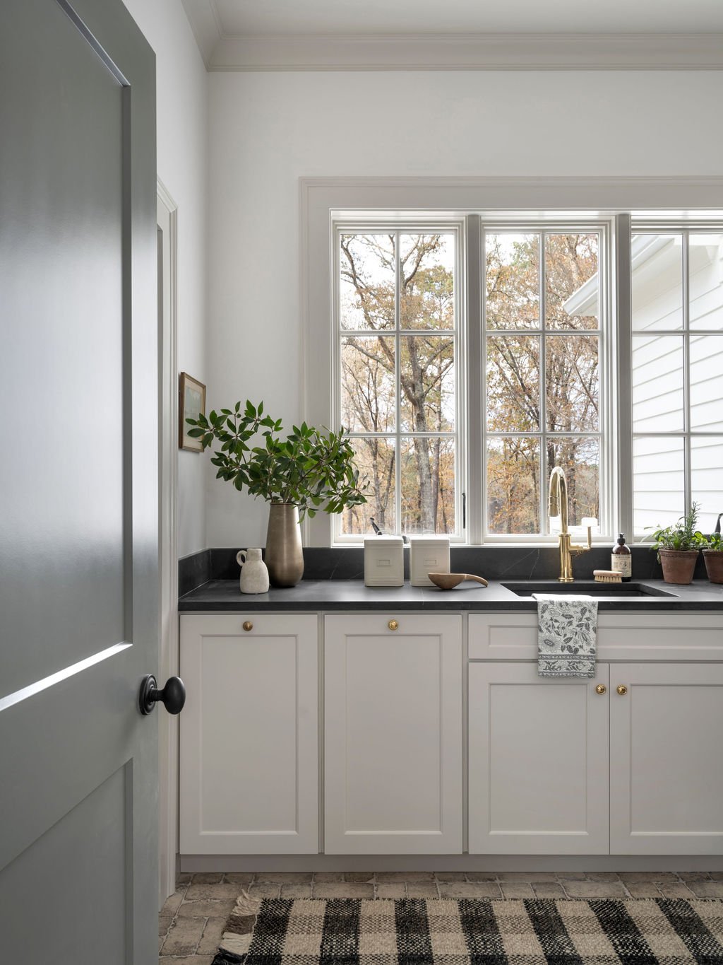 A kitchen with white cabinets, black countertop, and a large window showing trees outside. Decorations include a potted plant, small ceramic vases, and a soap dispenser.