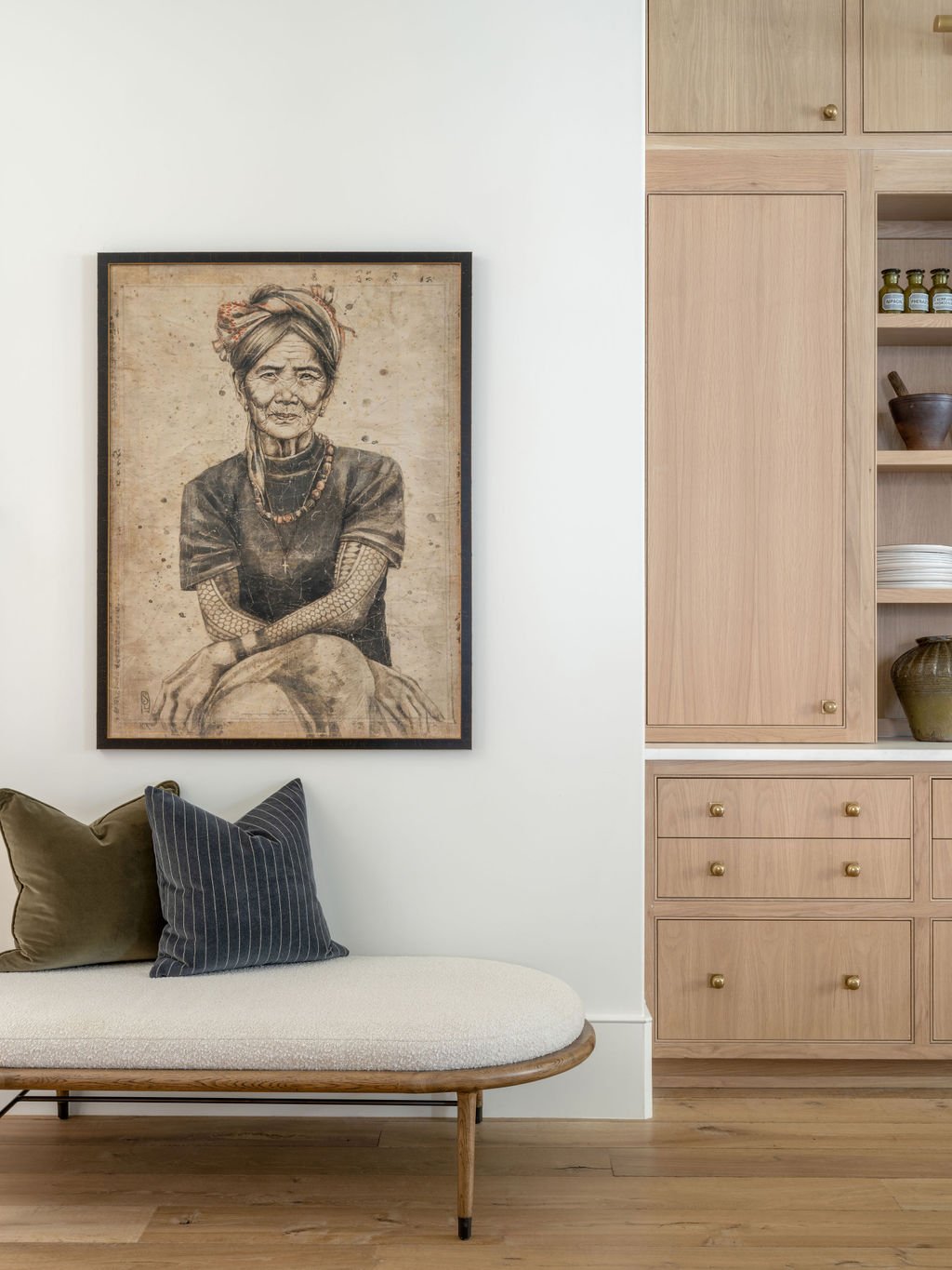 Interior of a home with a framed black and white portrait of an elderly woman hanging on the white wall. Below the picture is a beige cushioned bench with three pillows, one green and two dark blue with stripes. To the right, there is a light wooden 