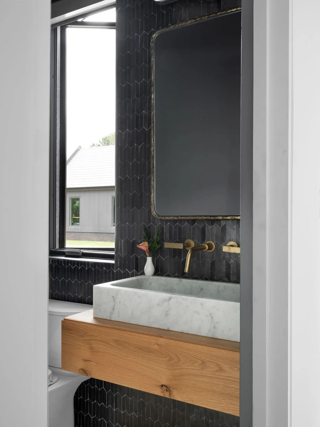 Modern bathroom with black hex tile wall, marble sink, brass faucet and mirror, small plant in a white vase, and window showing a house outside.
