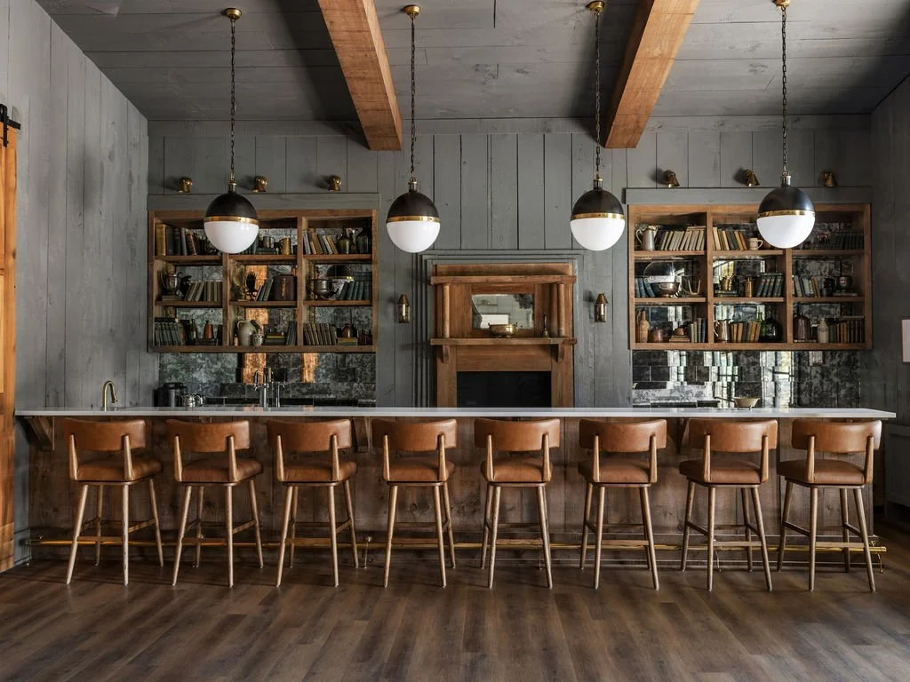 A rustic kitchen with a long wooden bar counter and eight matching wooden bar stools. Behind the counter are open wooden shelves filled with books and decorative items, a fireplace with a mirror above it, and pendant lights hanging from the ceiling. 