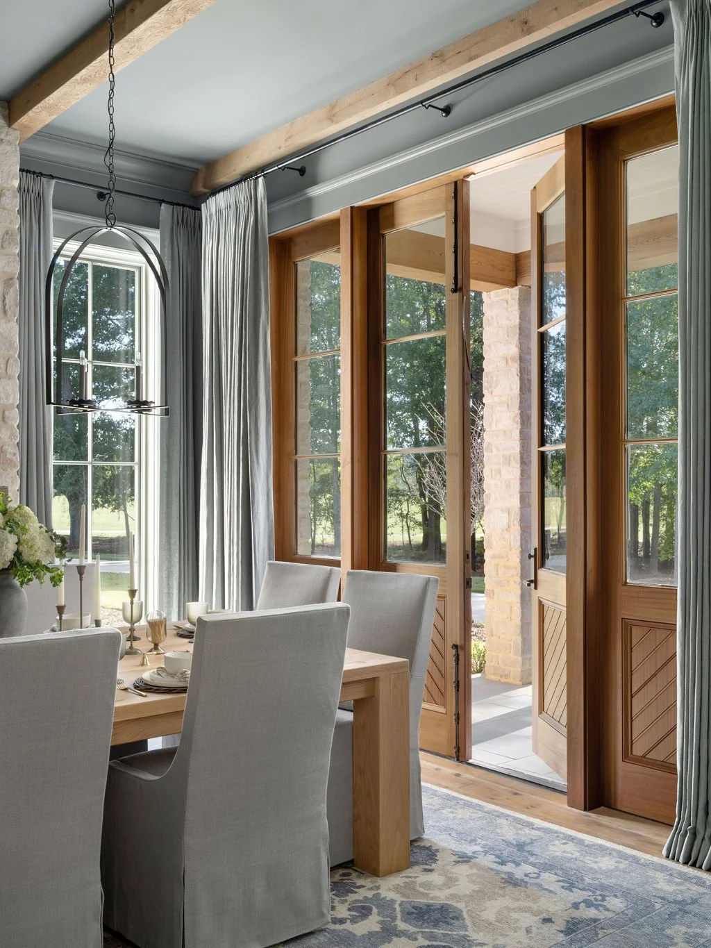 Dining room with a wooden table and six upholstered chairs, large windows with wooden frames, and an open door leading outside, with curtains and a modern chandelier hanging from the ceiling.