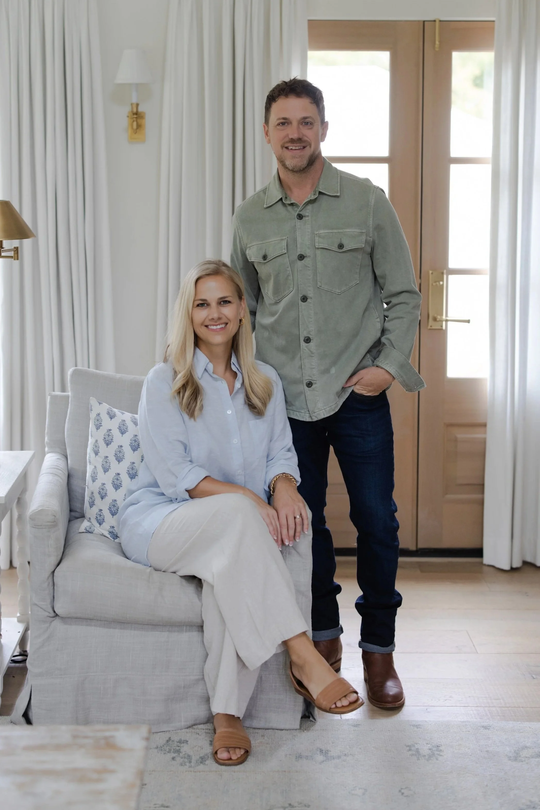 A smiling woman sitting on a light-colored armchair with a man standing behind her in a well-lit room with white curtains and a wooden door.
