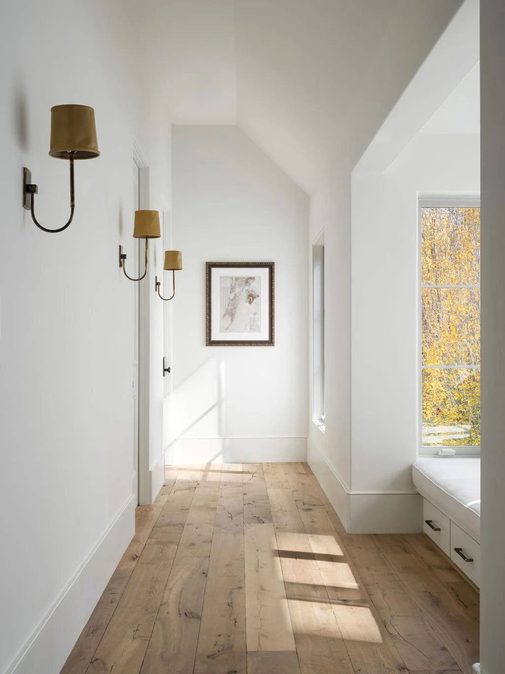 Bright hallway with white walls, hardwood floor, framed art on the wall, and a window with a view of autumn trees.