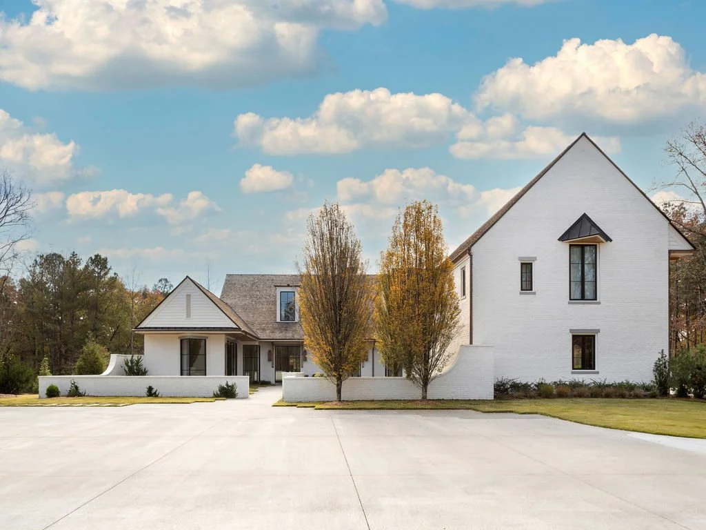 A large white house with a sloped roof and several tall windows, surrounded by trees with autumn foliage, under a partly cloudy sky.