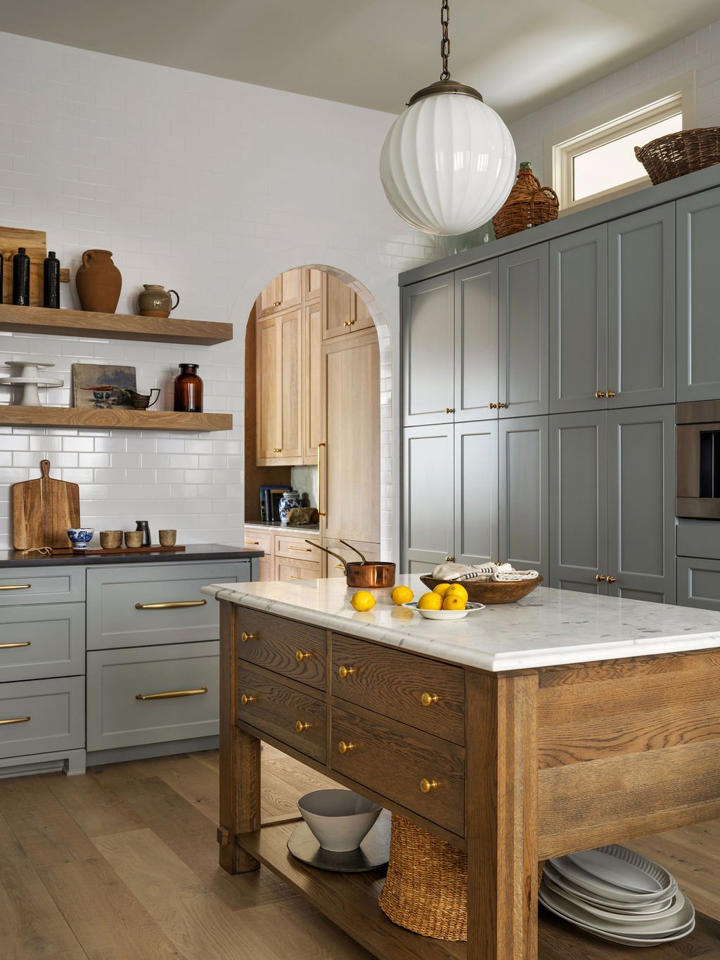 A modern kitchen with gray cabinets, a white marble island, wooden shelves with pottery, and fresh lemons on the island.