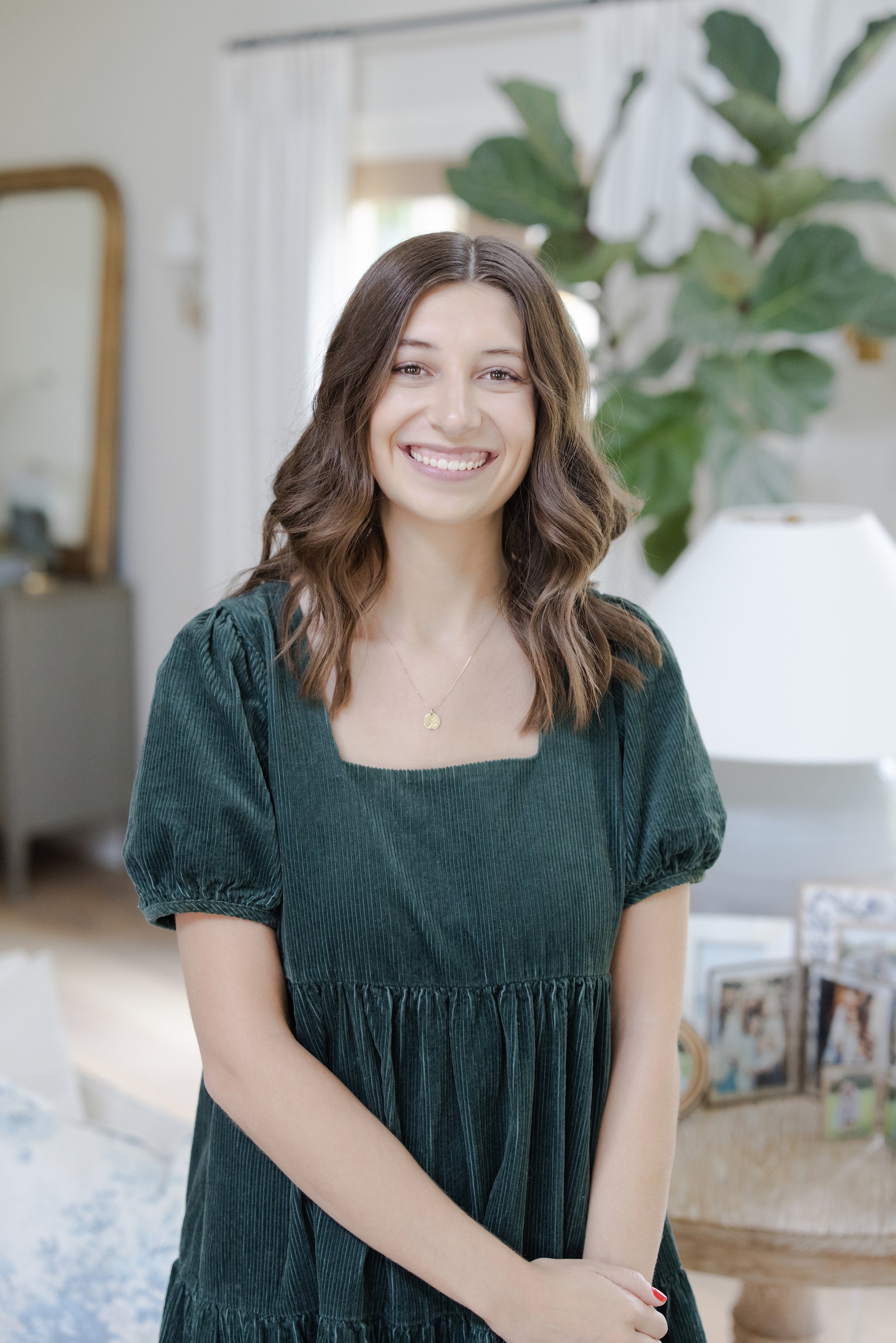 A young woman with shoulder-length brown hair smiling, wearing a dark green dress with puffed short sleeves, indoors in a well-lit room with a large leafy plant and framed photographs in the background.