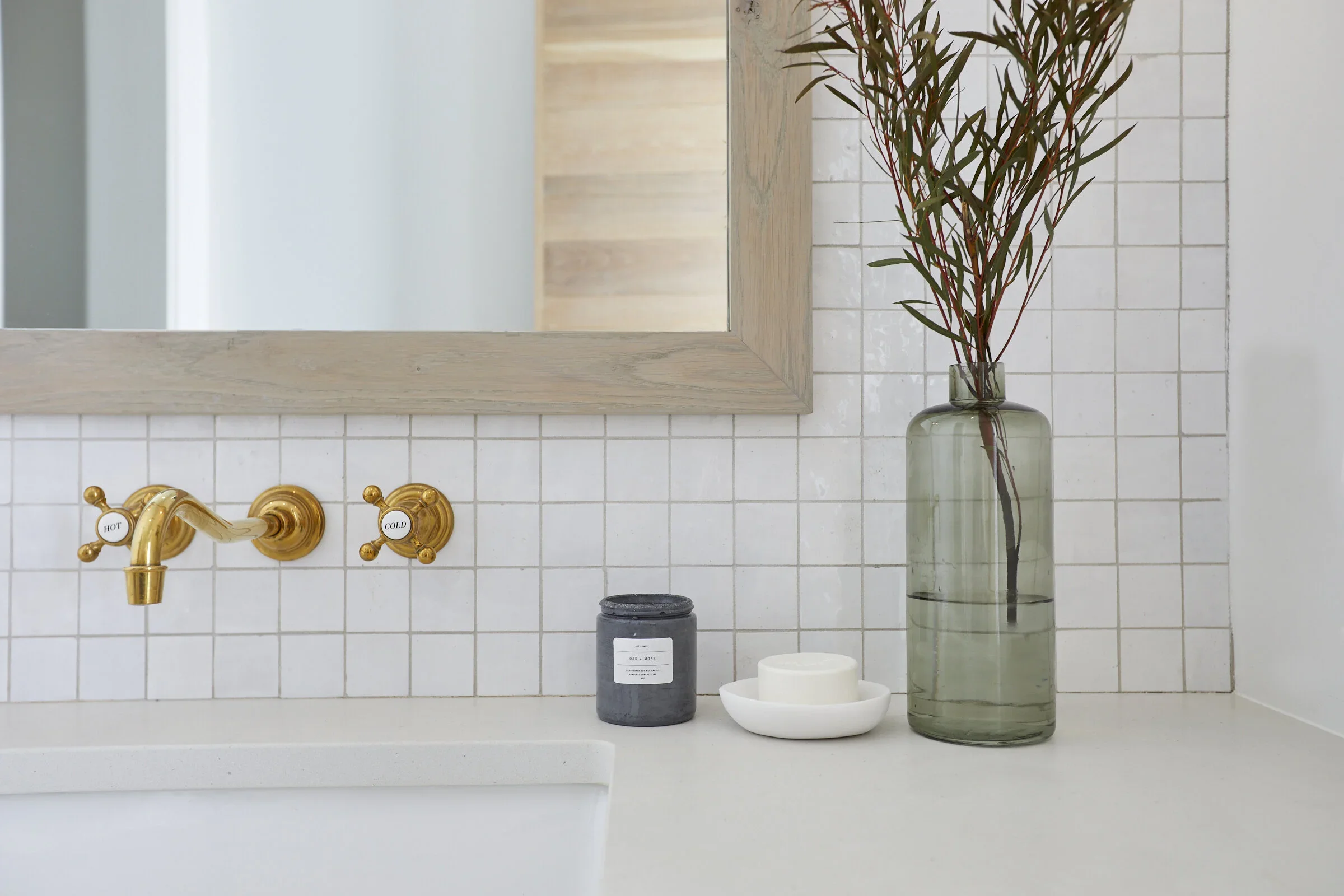 Bathroom sink area with gold faucet and handles, a vase with tall green and reddish leaves, a black candle, and a white candle in a small white dish on a beige countertop.