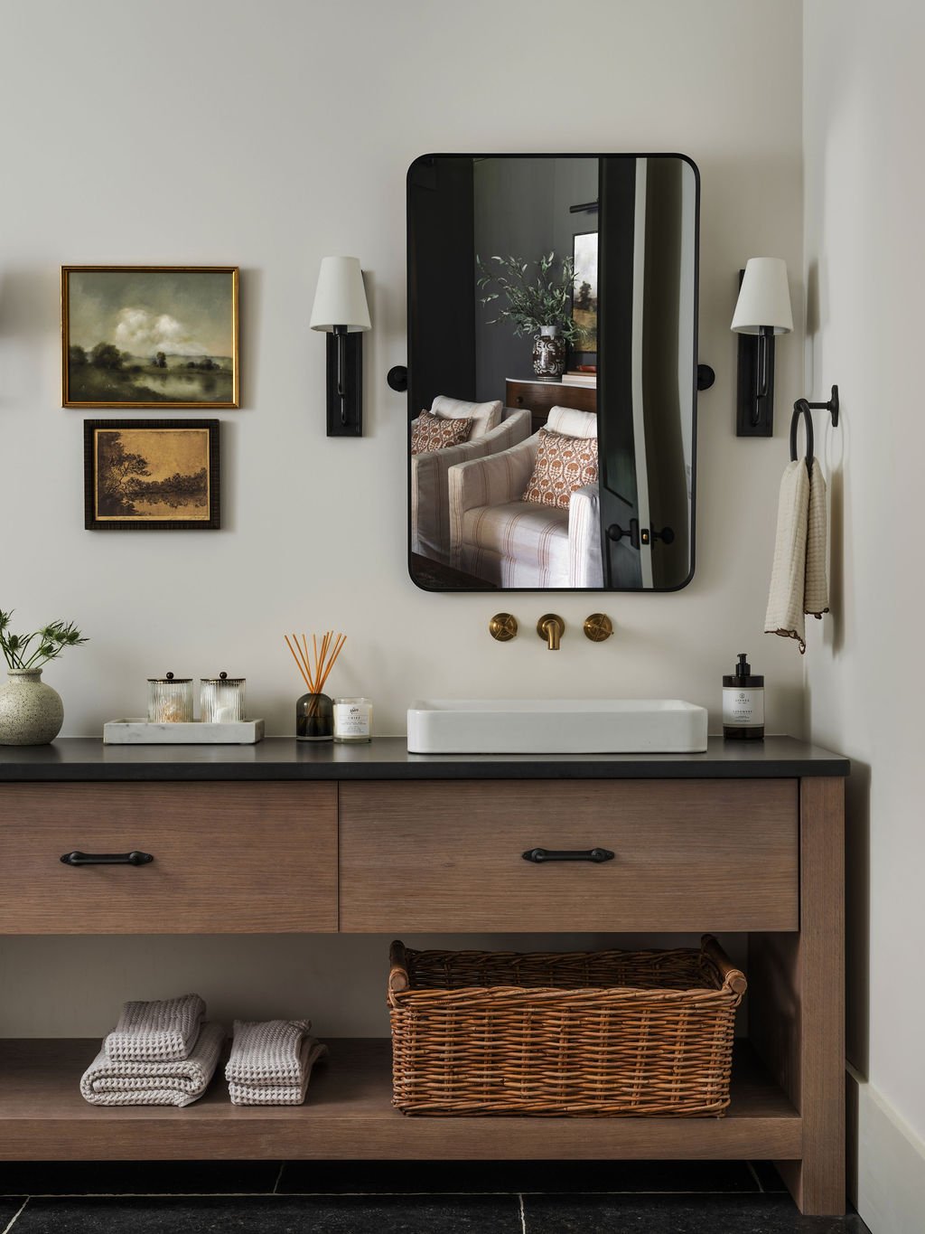 Bathroom with a wooden vanity, white basin sink, black countertop, and a large black-framed mirror. Decor includes wall-mounted lamps, framed art, a towel, toiletries, candles, a diffuser, and a wicker basket underneath.