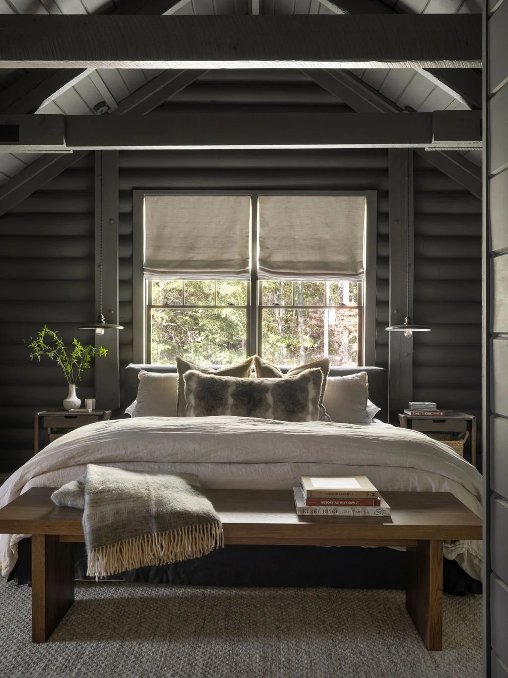 Cozy bedroom with a bed, window with beige blinds, pillows, books on a wooden bench, and a side table with a plant and decor.