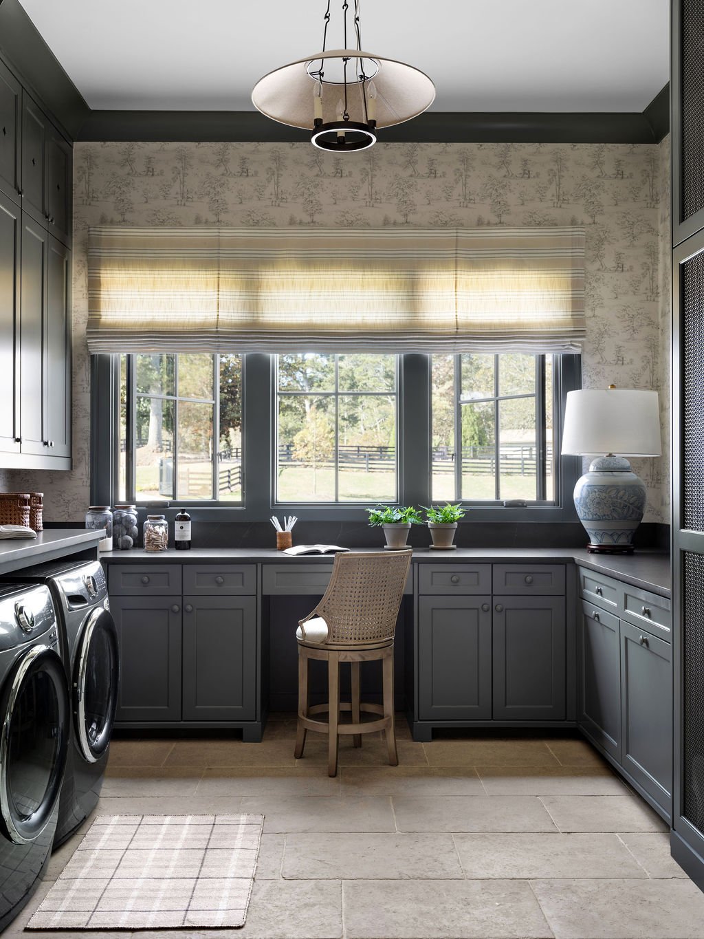 Laundry room with gray cabinets, large window with striped curtain, washer and dryer, and a desk with a chair, lamp, and decorative items.