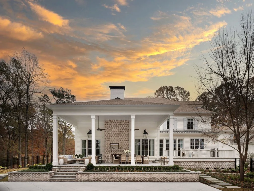 Exterior view of a large white two-story house with a covered porch and brick accents, set against a sunset sky with fall trees in the background.