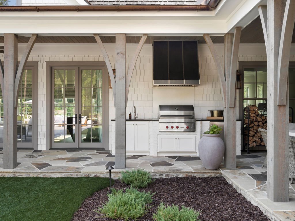 Outdoor kitchen with a stainless steel grill, white cabinetry, large potted plant, wood storage, and French doors leading inside, surrounded by a covered patio with stone flooring and a landscaped yard.