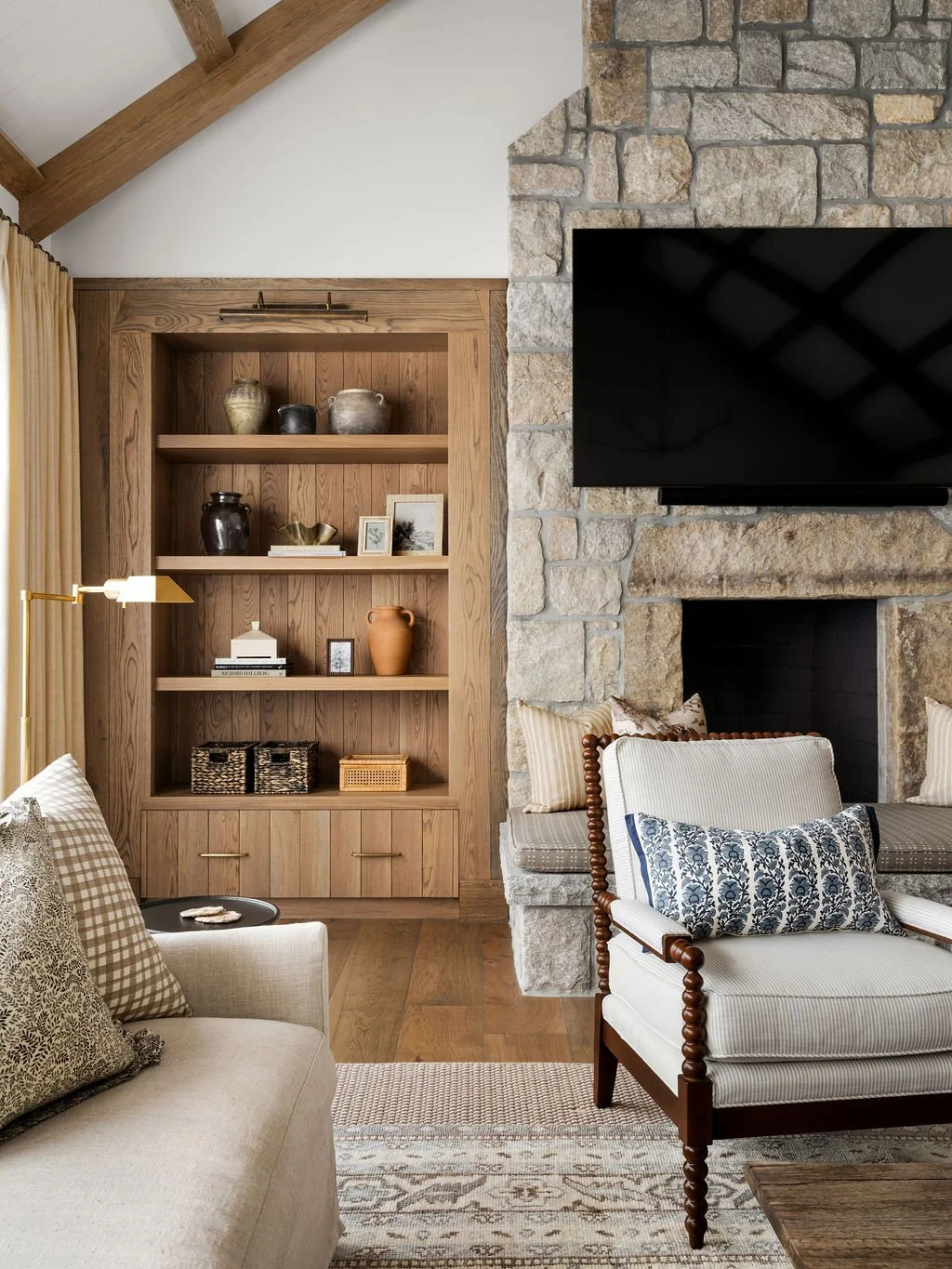 Living room with wooden and stone fireplace, black TV, beige and patterned furniture, and decorative items on a built-in wooden shelf.