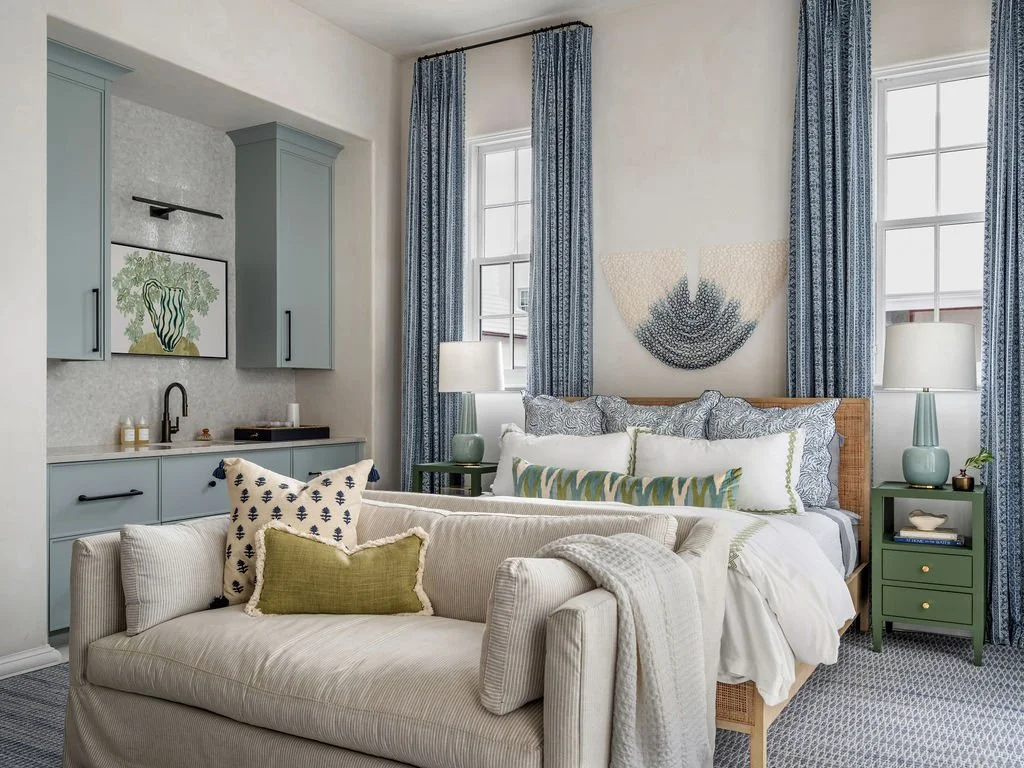 A bedroom with a bed, pillows, nightstands with lamps, curtains, and artwork, featuring light blue, white, and green decor.