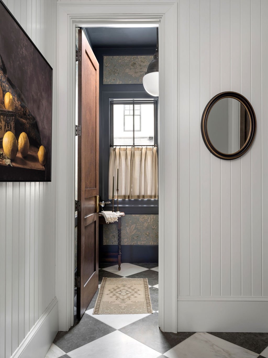 Interior view of a doorway leading to a small room with floral wallpaper, a window with a curtain, and a hanging light fixture. The floor has a patterned tile design with a small rug near the doorway.