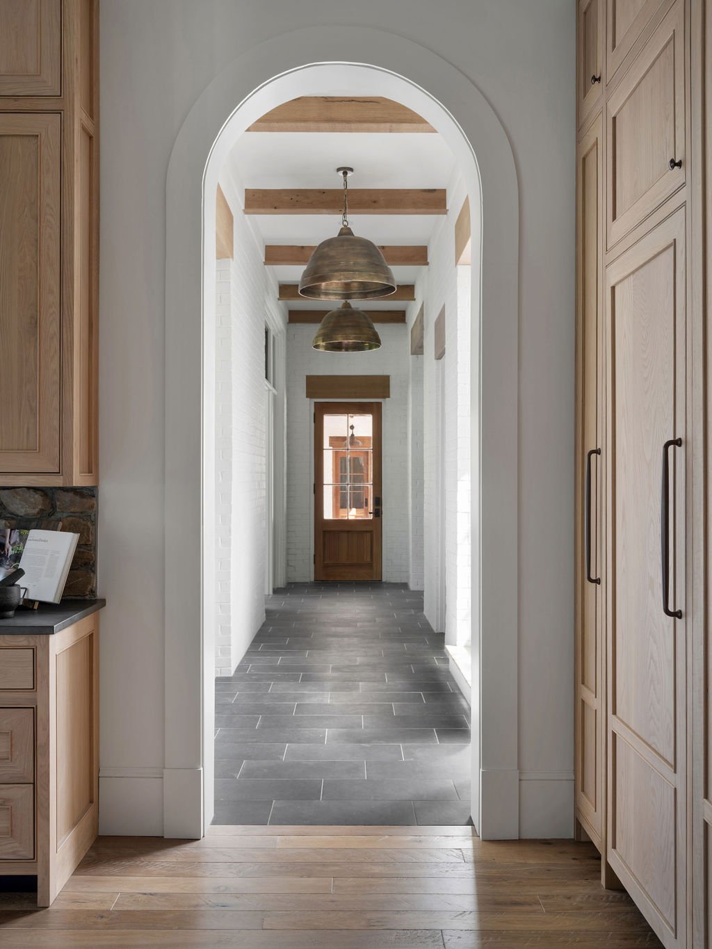 View through an arched doorway into a hallway with wooden ceiling beams and hanging bronze pendant lights. The hallway has white brick walls and dark grey tile flooring, leading to a wooden door at the end.