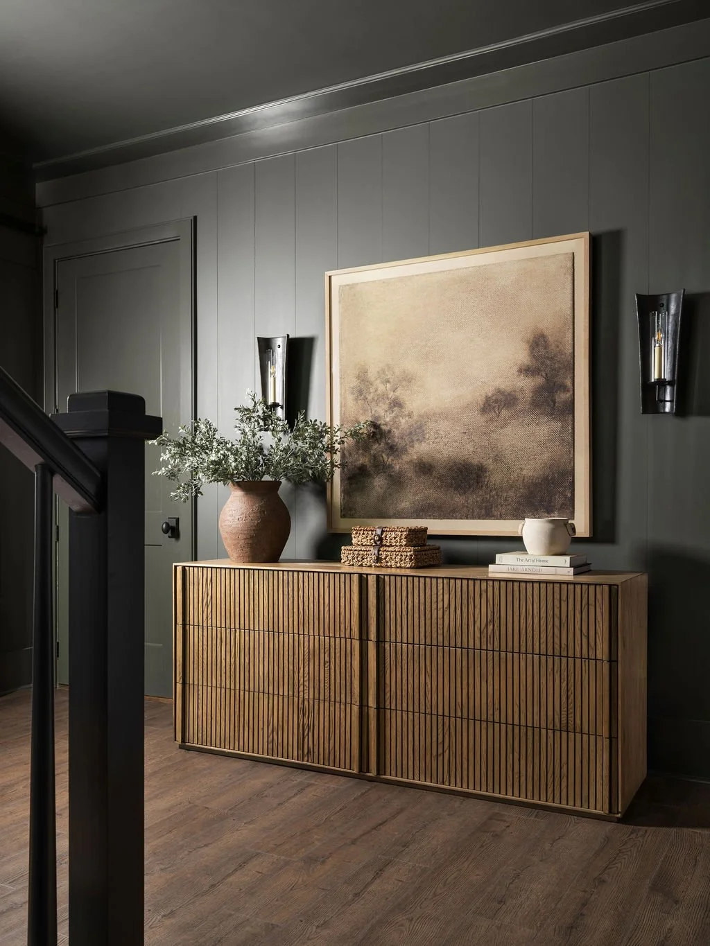Modern interior hallway with a wooden sideboard, large framed artwork, potted plant, and decorative items, gray paneled walls, wall-mounted sconces, and hardwood flooring.