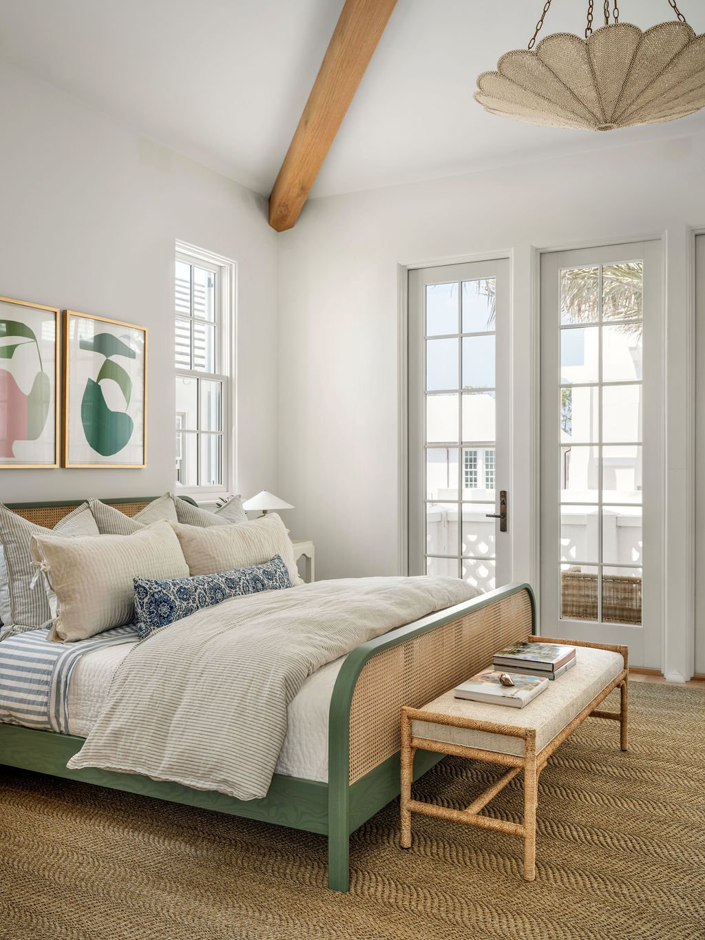 A bedroom with a white bed, multiple pillows, and framed abstract artwork above the headboard, next to a small white nightstand with a lamp. French doors lead outside, and a woven bench with books is at the foot of the bed. The ceiling has a wooden b