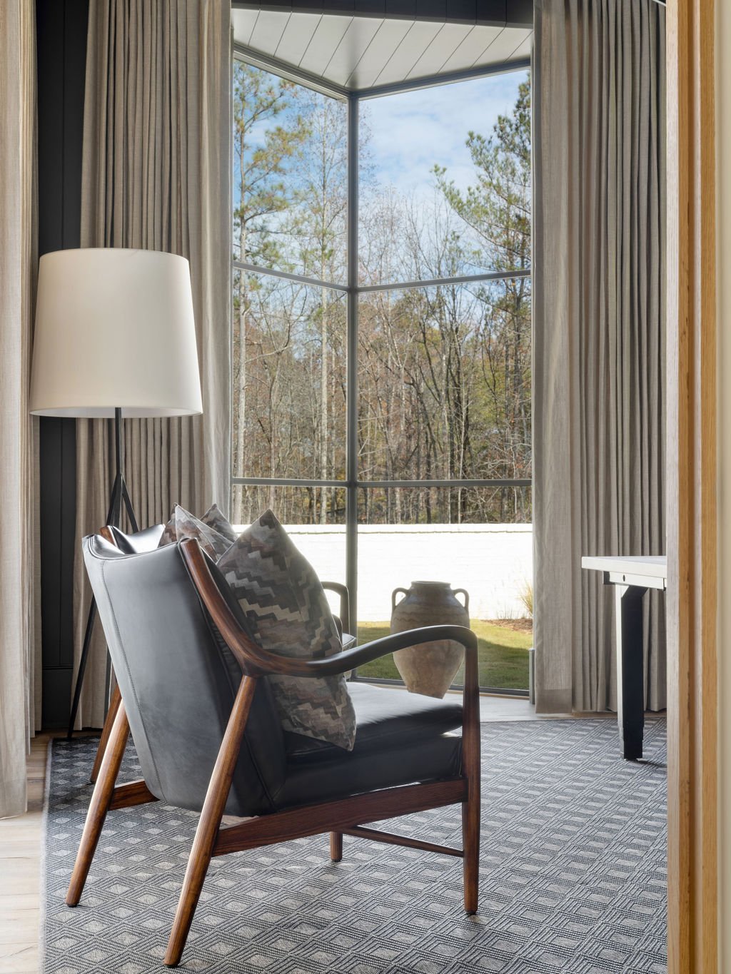 View from inside a room looking out a large corner window revealing a yard with trees and a vase outside. Inside the room, there is a black armchair with wooden arms and patterned pillows, a tall white floor lamp, beige curtains, and a small black ta