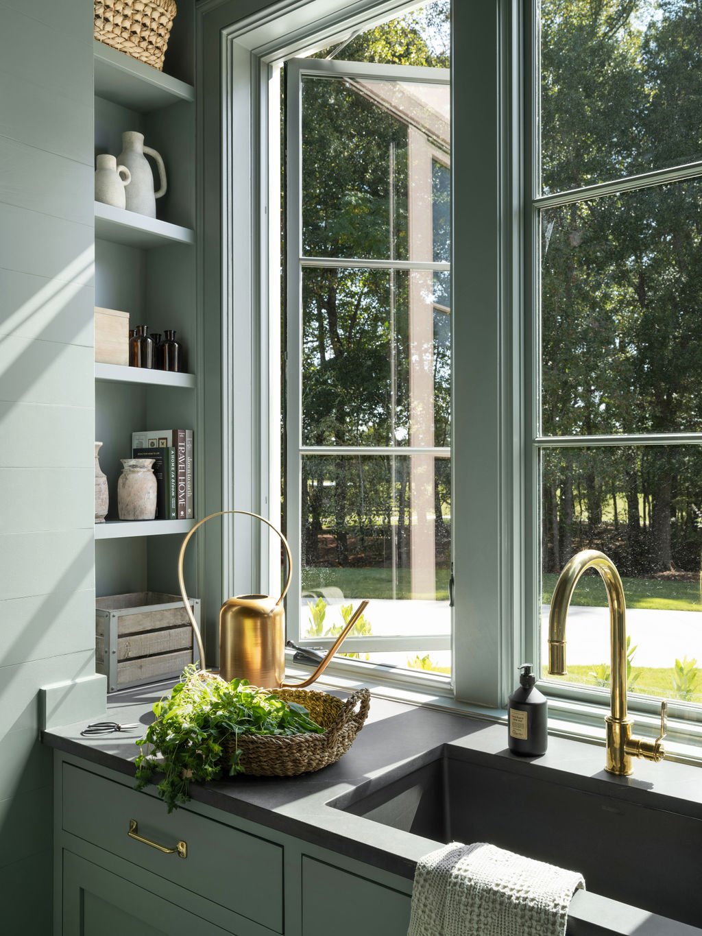 A kitchen window overlooks a garden with trees. The kitchen has light green cabinets, open shelves with decorative items, and a gray countertop with a basket of greens, a watering can, and a soap dispenser near a black sink with a gold faucet.